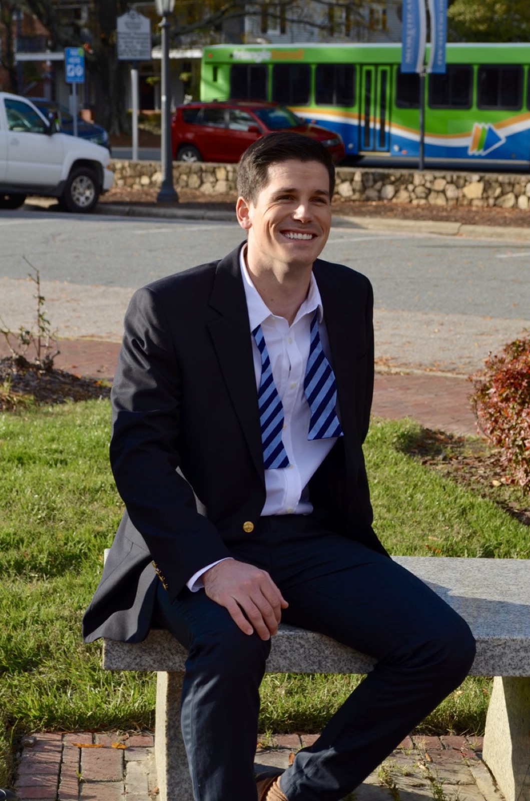 A man seated on a stone bench in a navy one-piece Suitsy business suit with a blue striped necktie, a bus passing behind him.