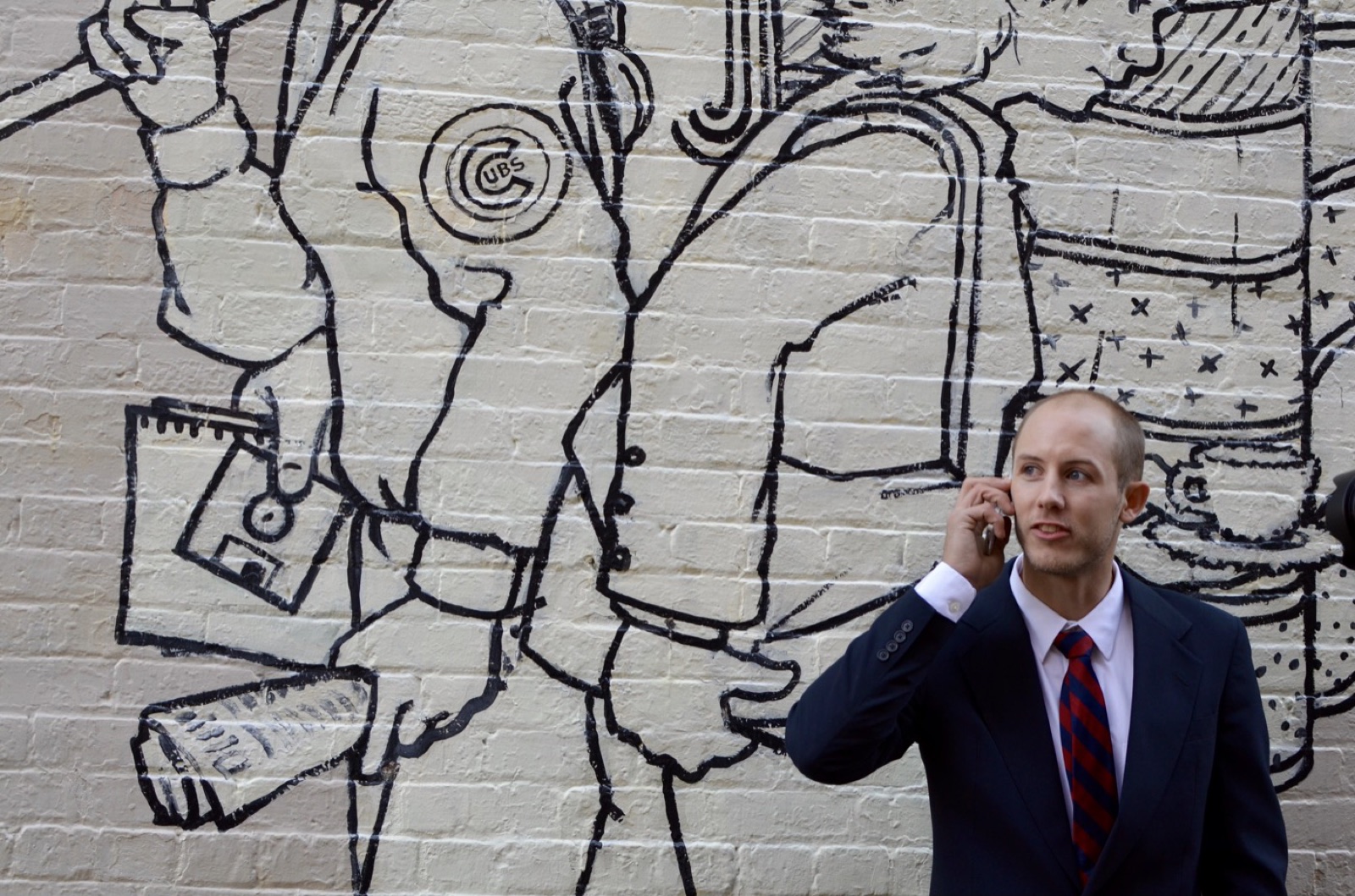 A man in a navy one-piece Suitsy business suit with a red and navy striped tie, taking a phone call in front of a large black-and-white line-drawing mural on a painted brick wall.