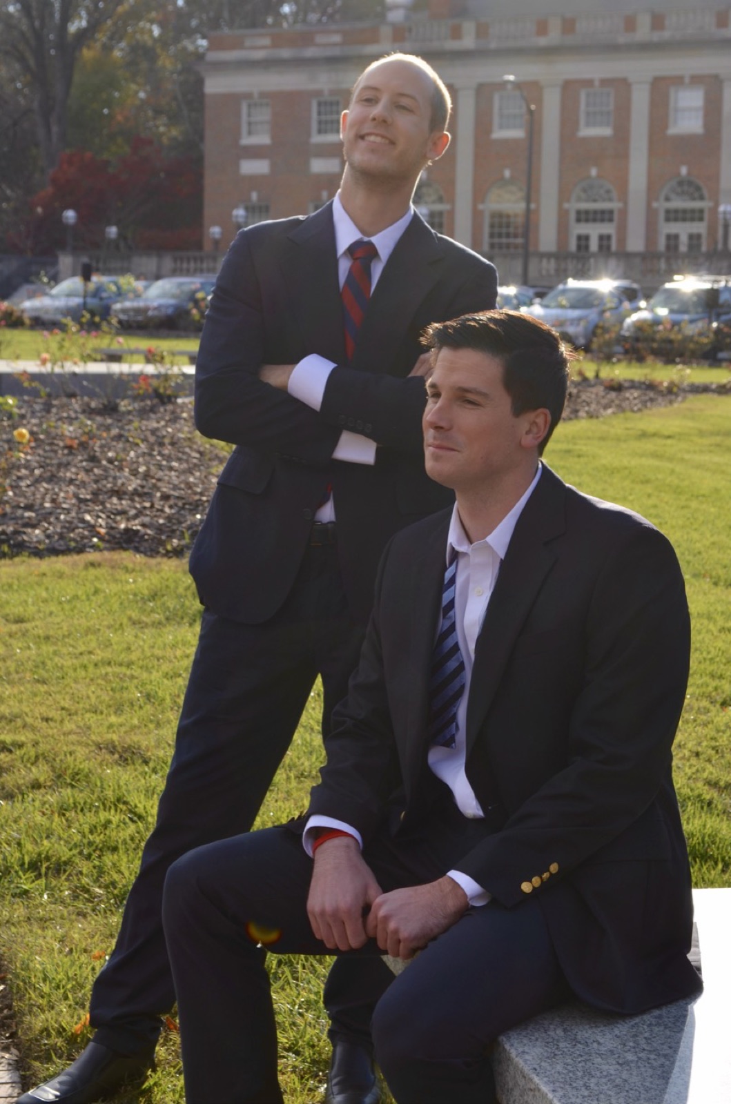 The same two men, one standing with arms folded and one seated on a stone bench, both in dark one-piece Suitsy business suits.