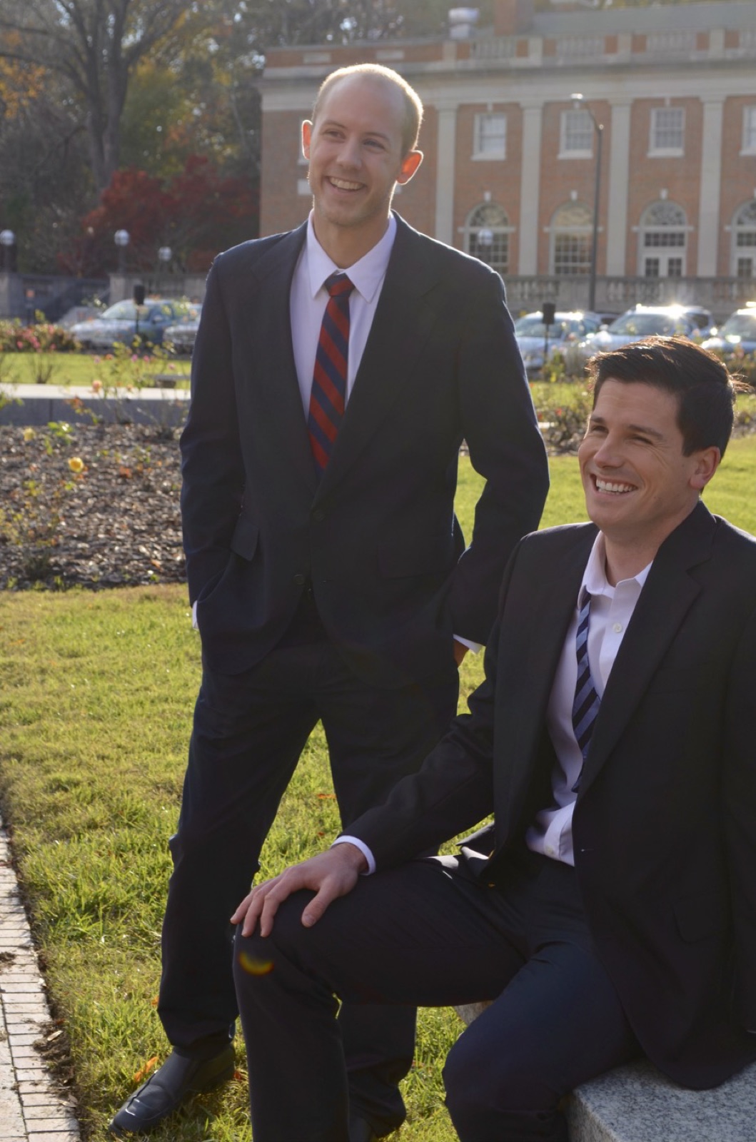 Two men outside a brick building in autumn, both wearing dark one-piece Suitsy business suits with white shirts and striped neckties, laughing.